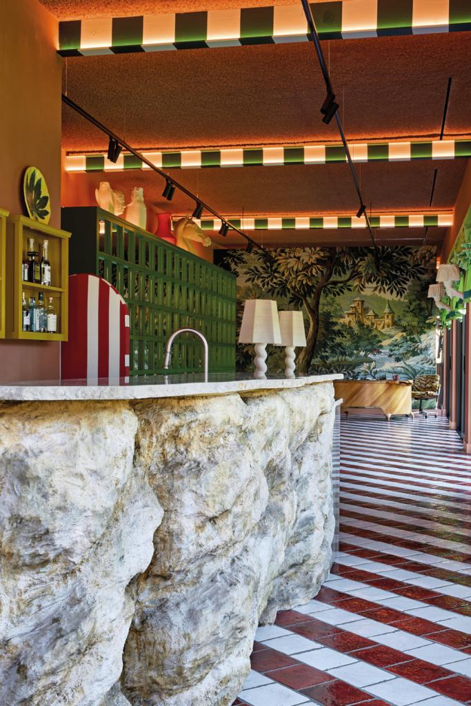 Keuken en thuiskantoor in Puurs. ‘In plaats van de stalen balken te verstoppen onder een verlaagd plafond, voorzagen we ze van indirecte verlichting en een Italiaans heraldisch streepdessin gecombineerd met een jarentachtigkast en een Vlaams wandtapijt. De “kruiding” zit ‘m in de rots onder het aanrecht en de bureautafel in de vorm van een leeuw.’