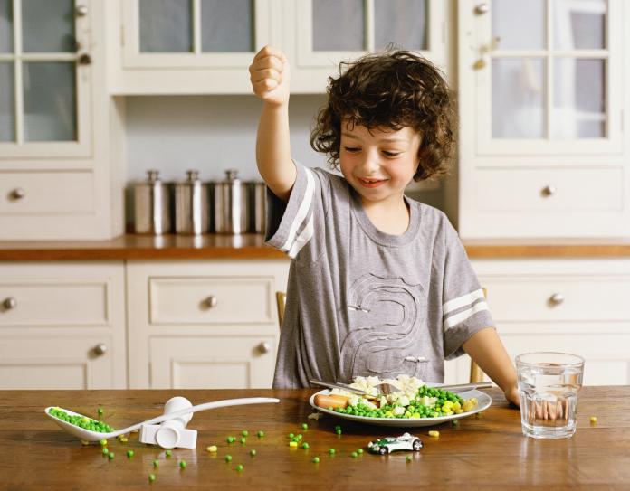 Boy (5-7) with fist raised over spoon handle, peas on table