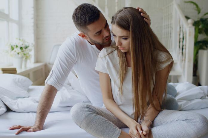 Depressed young woman sitting in bed and crying while her boylfriend come and embrace her and kiss in bedroom at home