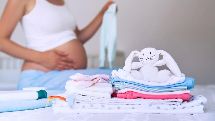 Pile of baby clothes, necessities and pregnant woman on bed in home interior of bedroom.