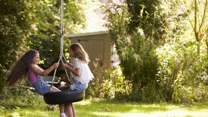 Two Girls Playing Together On Tire Swing In Garden