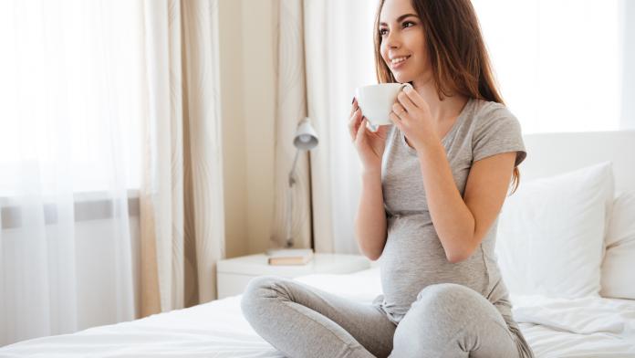 Smiling pregnant woman drinking coffee on bed at home