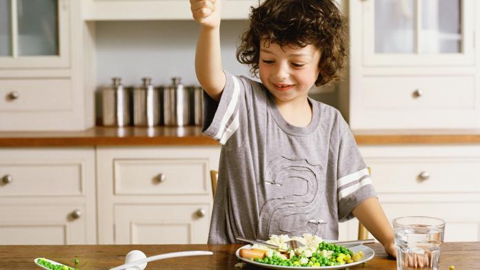 Boy (5-7) with fist raised over spoon handle, peas on table