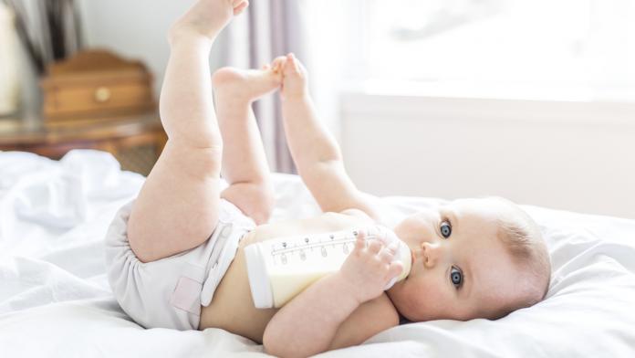 Pretty baby girl drinks water from bottle lying on bed. Child weared diaper in nursery room