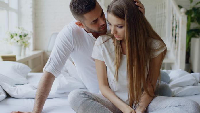 Depressed young woman sitting in bed and crying while her boylfriend come and embrace her and kiss in bedroom at home