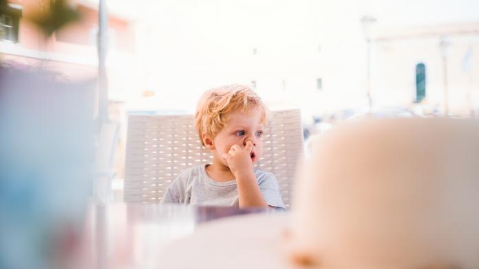 Small boy sitting in outdoor restaurant on summer holiday, picking nose. neuspeuteren