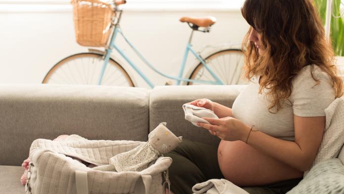 Pregnant Mid Adult Woman Looking Baby Clothing While Sitting On Sofa At Home zwanger zijn en bevallen corona