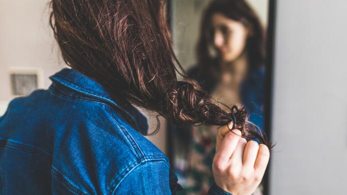 Young woman standing in front of mirror, pulling hair-band from hair, rear view