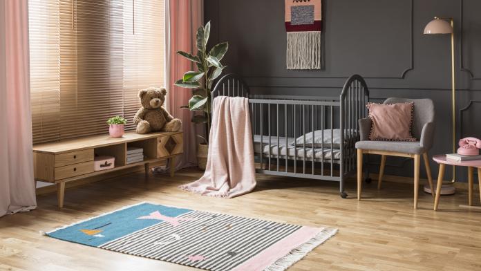 Real photo of a baby crib standing between a low cupboard and an armchair, lamp and stool in child's room interior with wooden floor and grey walls with moldings