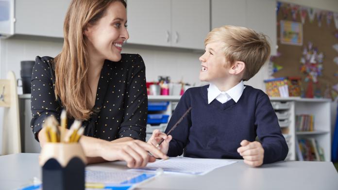 Young female primary school teacher and schoolboy sitting at a table working one on one, looking at each other smiling, front view, close up oudercontact kindcontact kindgesprek