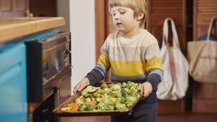 Helpful young boy loading a baking tray full of organic vegan vegetables into oven vegetarisch eten kinderen