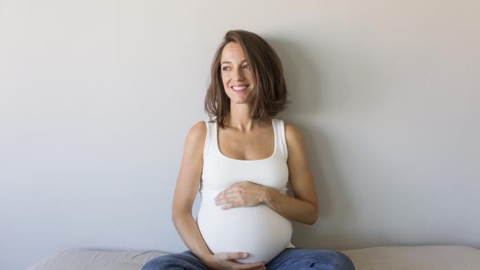 Smiling Pregnant Woman Touching Belly While Sitting On Bed At Home
