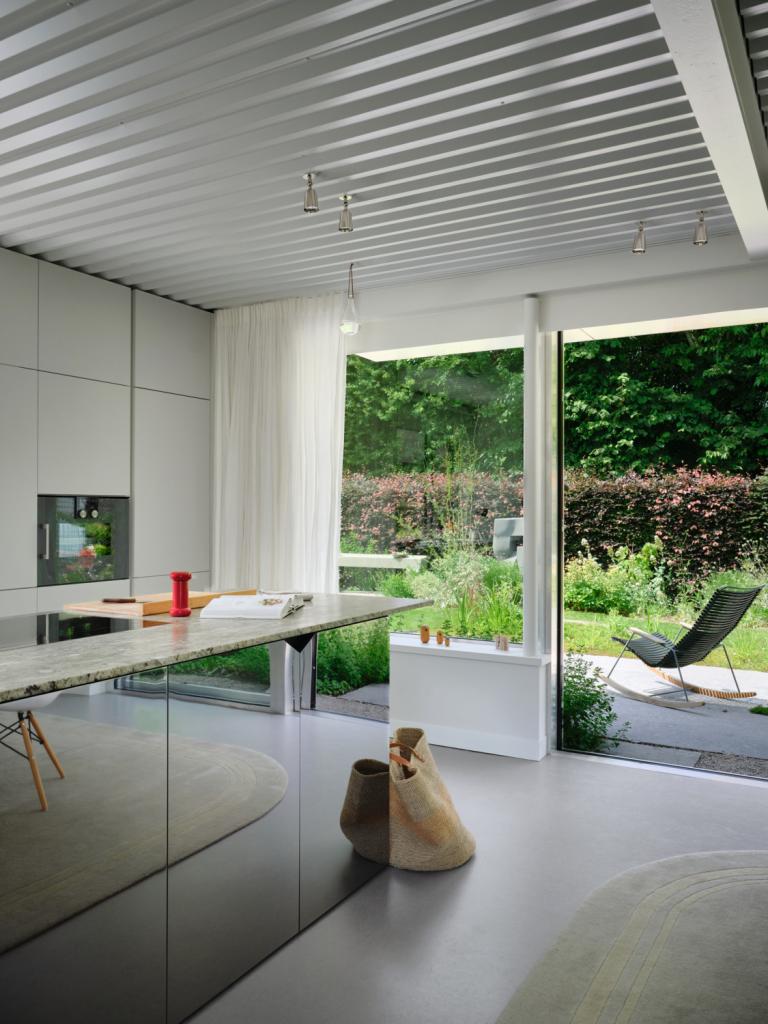 Witte keuken met spiegelei kookeiland. Zowel buiten als binnen worden spiegeloppervlakken ingezet om de connectie met groen te versterken. Het beeld in de tuin werd gemaakt door bevriend kunstenaar Jean-Luc Verpoucke.