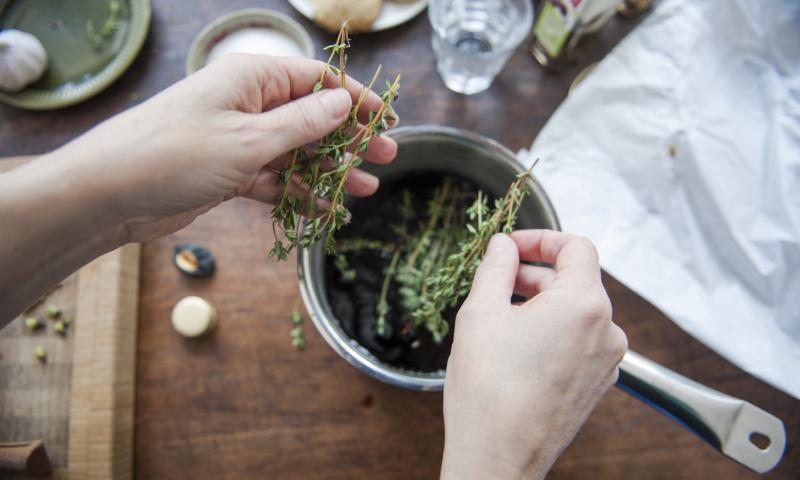 Woman adding fresh thyme leaves to a cooking pot filled with balsamic vinegar, to make a dressing. First person perspective.