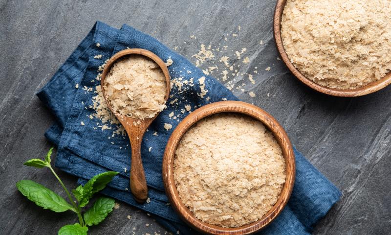 Dietary supplement, unfortified nutritional yeast flakes in a wooden bowl and scoop on stone backdrop