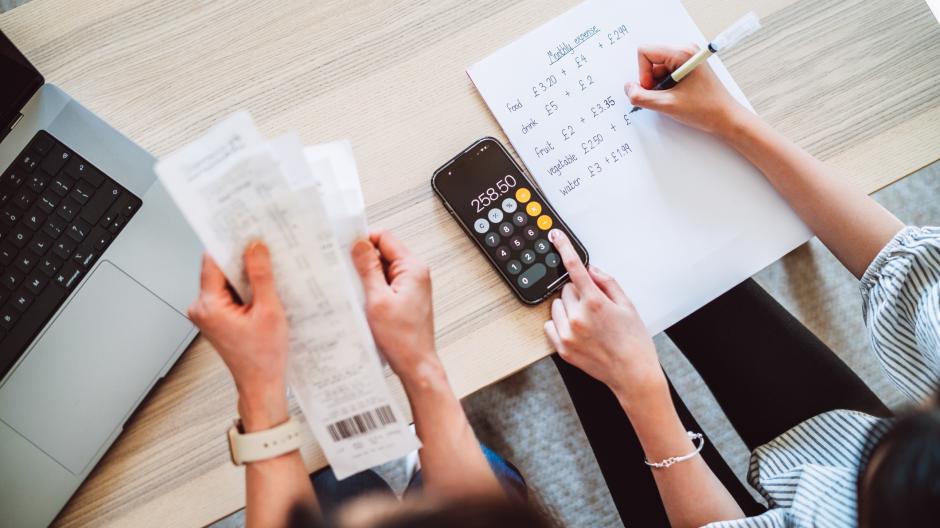 Een foto van twee personen die samen een gezinsbudget opstellen. De ene heeft enkele rekeningen vast, de andere maakt de berekeningen met een rekenmachine op de smartphone.