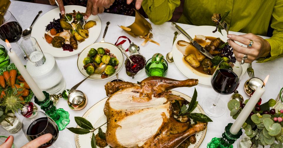 High Angle View Hands Of Friends Talking And Eating Traditional Christmas Turkey Dinner At Table With Red Wine And Candles