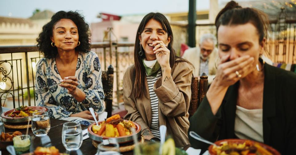 Medium shot of women laughing and savoring traditional Moroccan tajine at scenic rooftop eatery during holiday in Marrakech