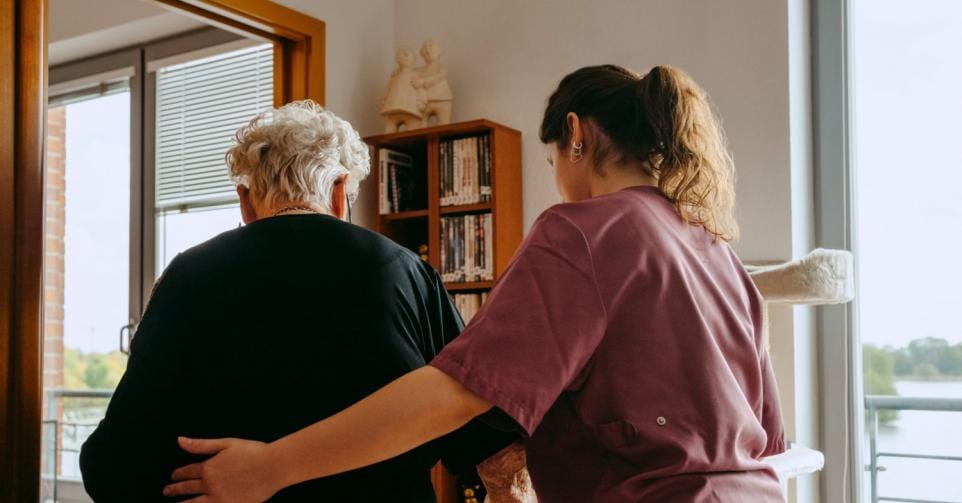 Een foto van een woonzorgcentrum waarop een oudere wordt begeleid door een medewerker.